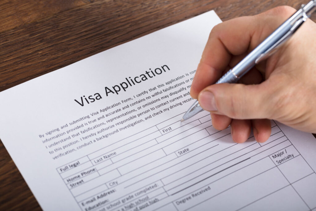 Close-up Of A Person Hand Filling Visa Application Form On Wooden Desk