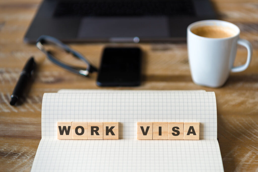 Closeup on notebook over wood table background, focus on wooden blocks with letters making Work Visa text. Concept image. Laptop, glasses, pen and mobile phone in defocused background